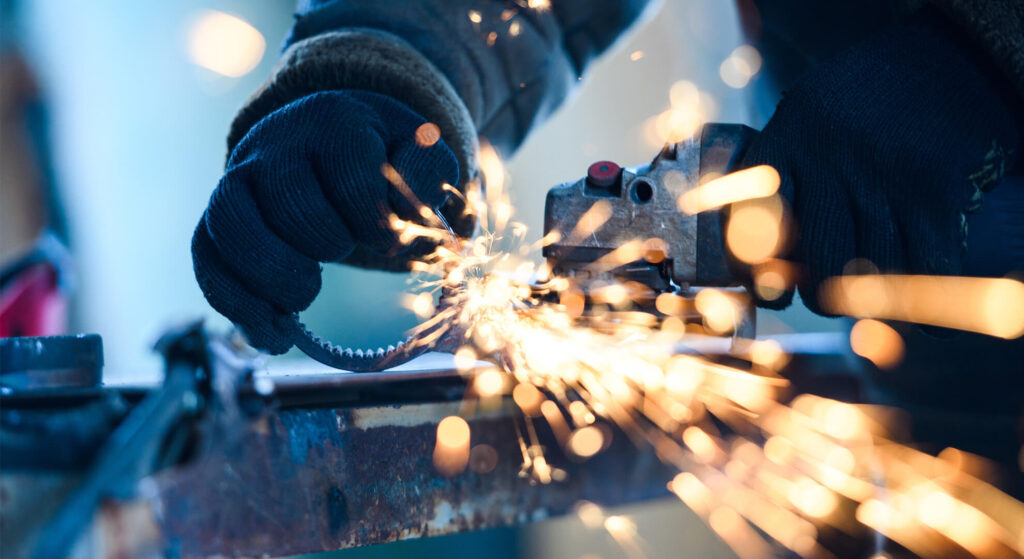 作業着と手袋を着用した職人が、グラインダーを使用して金属パーツを火花を散らしながら研磨・加工している手元の様子。