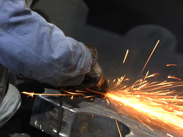 暗い工場内で、職人が電動工具を使って金属を研磨しているクローズアップ。作業箇所から鮮やかなオレンジ色の火花が長く尾を引いて飛び散っている。